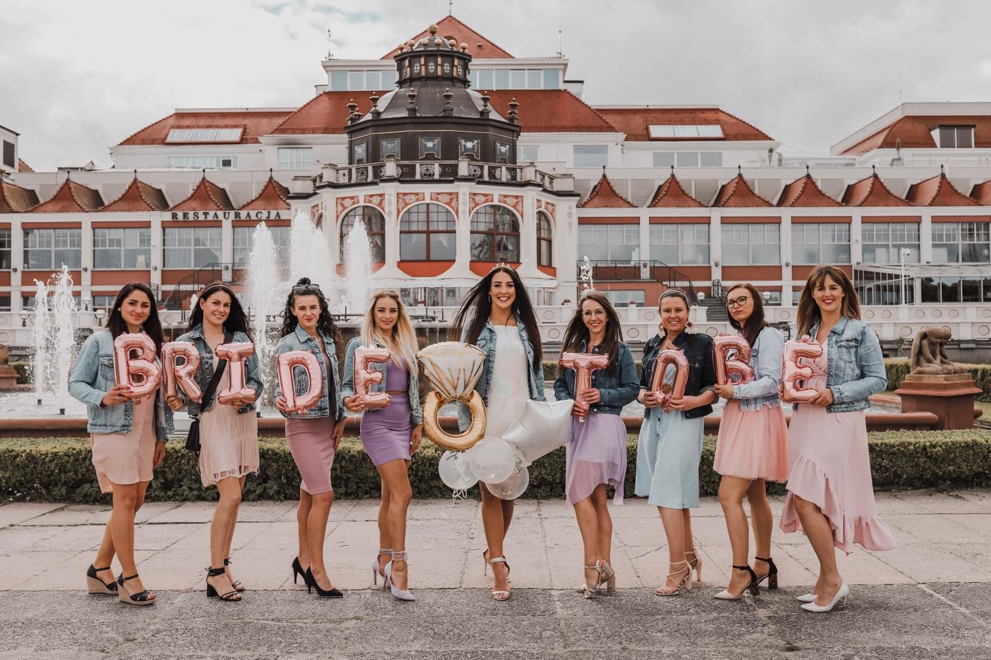 Grupa kobiet w pastelowych sukienkach i jeansowych kurtkach pozuje z balonami układającymi się w napis 'BRIDE TO BE' na tle fontanny i budynku restauracji w Sopocie.