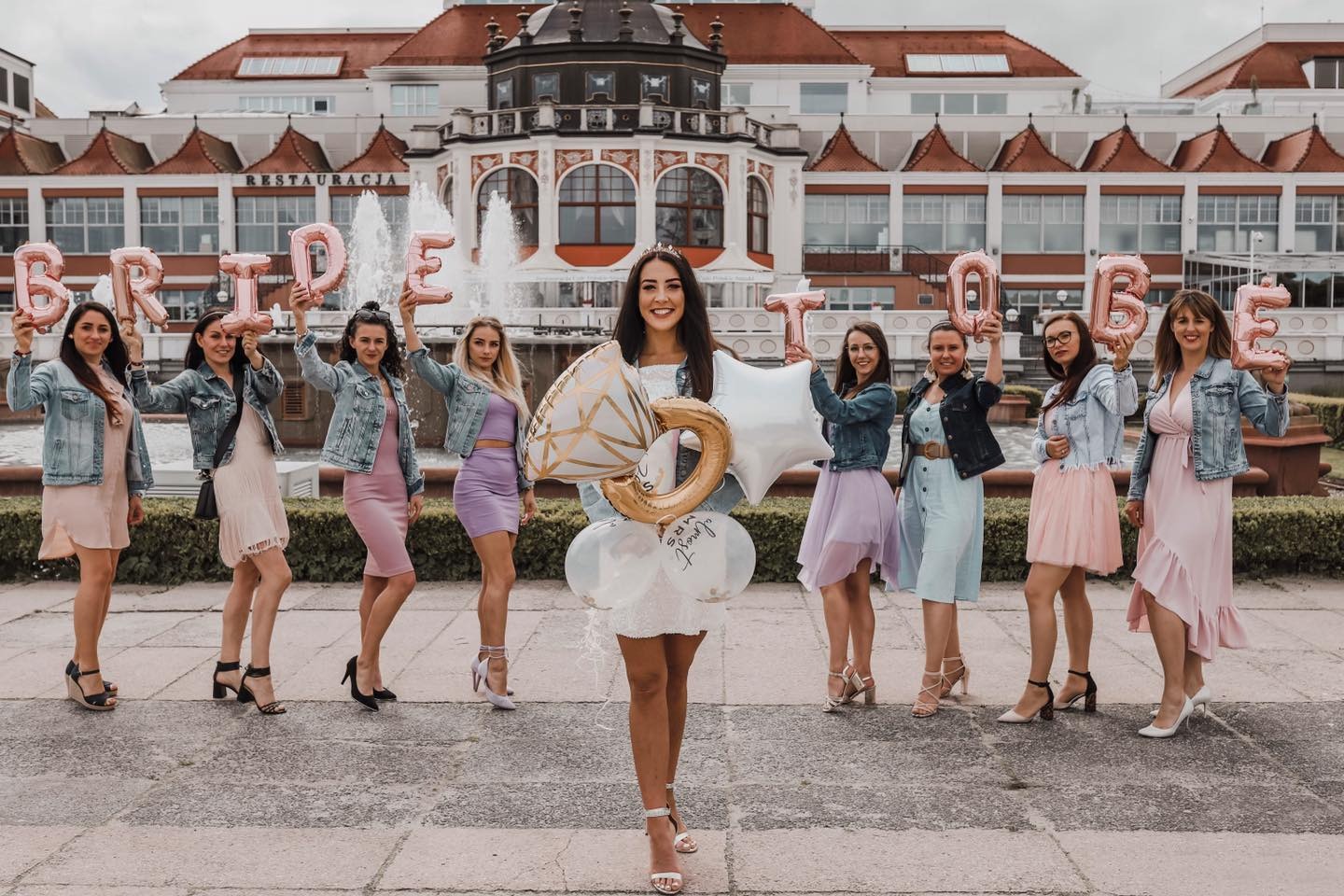Grupa kobiet świętuje wieczór panieński przed budynkiem z fontanną, trzymając balony w kształcie liter układających się w napis BRIDE TO BE oraz balony w kształcie diamentu i gwiazdy.