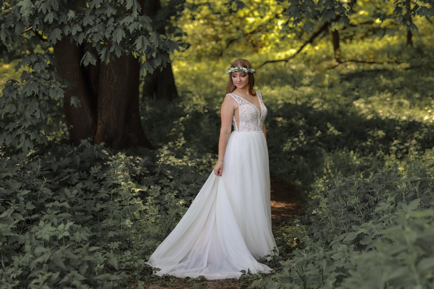 Kobieta w białej sukni z wiankiem na głowie pozuje w zaroślach leśnych, trzymając brzeg sukni.