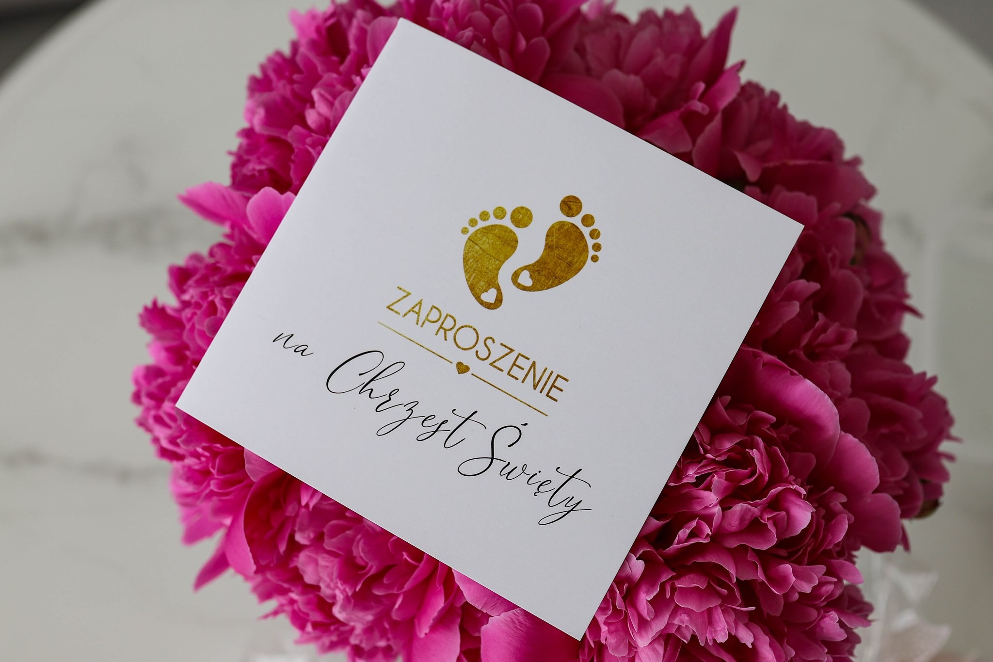 Eleganckie zaproszenie na chrzest z motywem złotych stópek niemowlęcych, umieszczone na tle bujnego bukietu różowych piwonii.