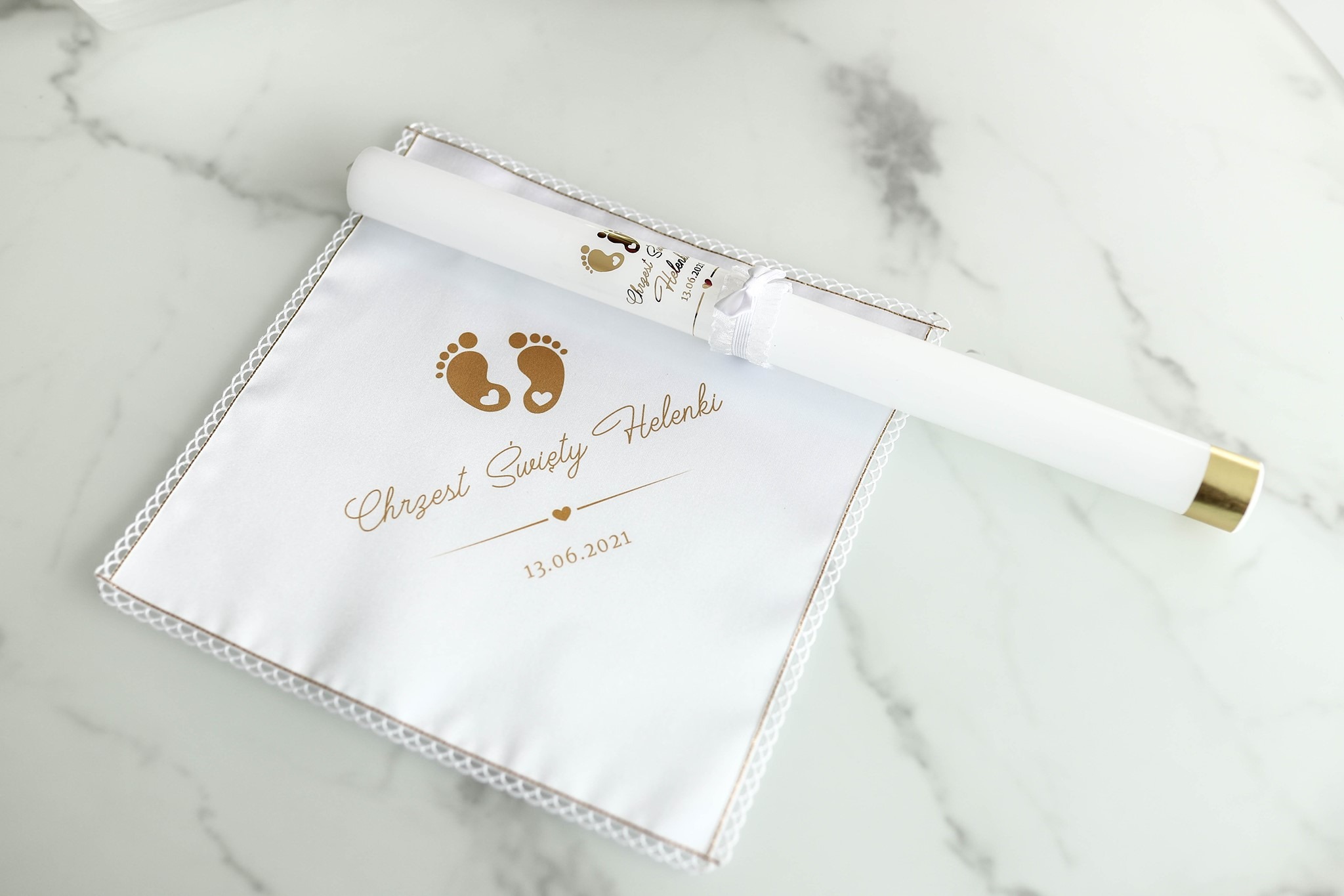 Świeca i haftowana chusteczka pamiątkowa z okazji Chrztu Świętego Helenki, z datą 13.06.2021 oraz złotymi odciskami stóp dziecka, leżące na marmurowym tle.