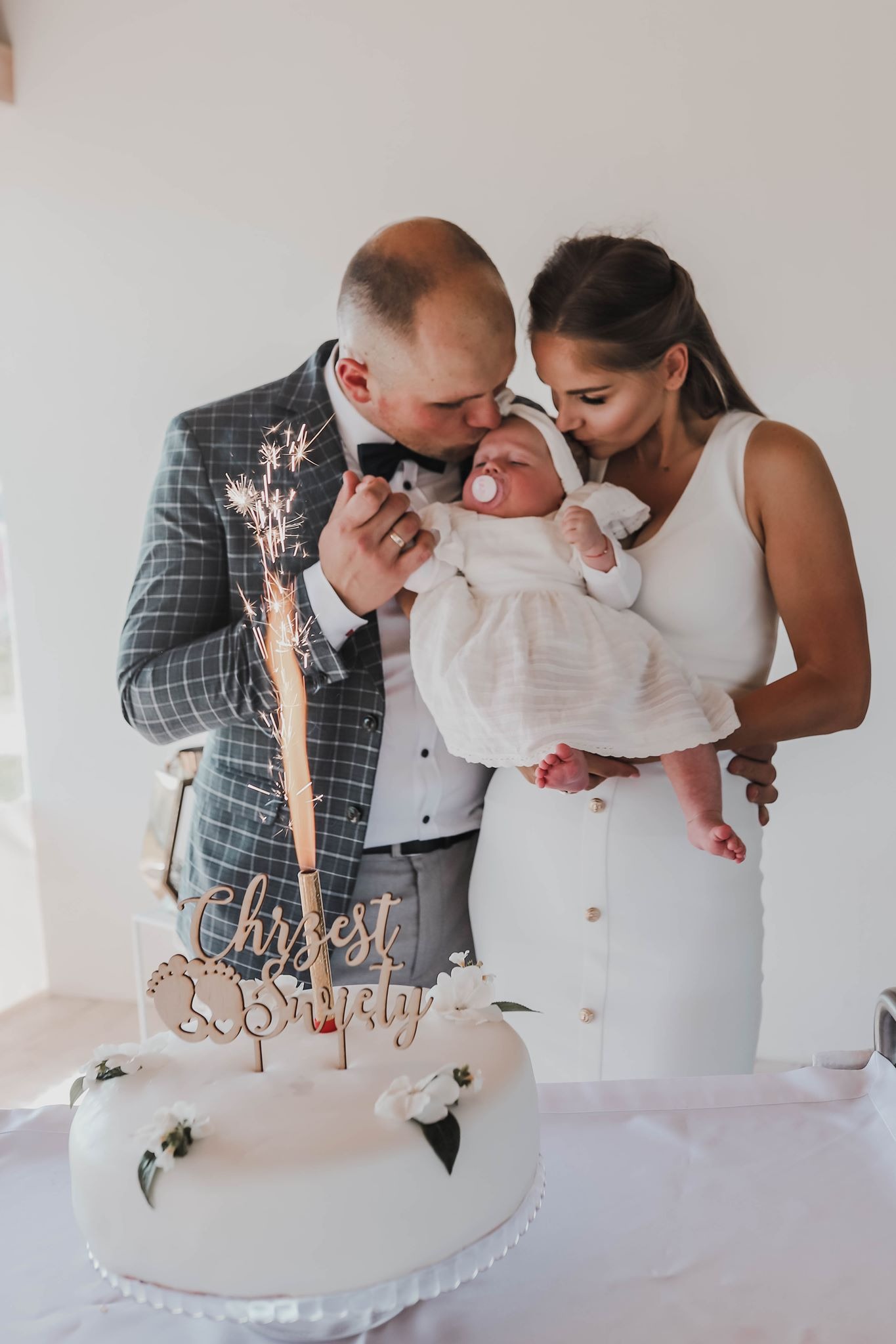 Rodzice całują śpiące niemowlę ubrane w białą sukienkę, na pierwszym planie tort z napisem 'Chrzest Święty' i płonącą zimną fontanną.