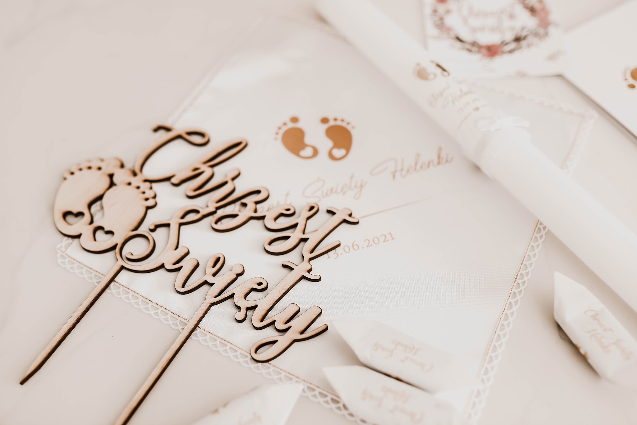 Dekoracje na chrzest: drewniane napisy 'Chrzest Święty' z motywem stóp dziecka, świeca z imieniem Helenki i datą 13.06.2021, całość na białej serwetce z koronką.