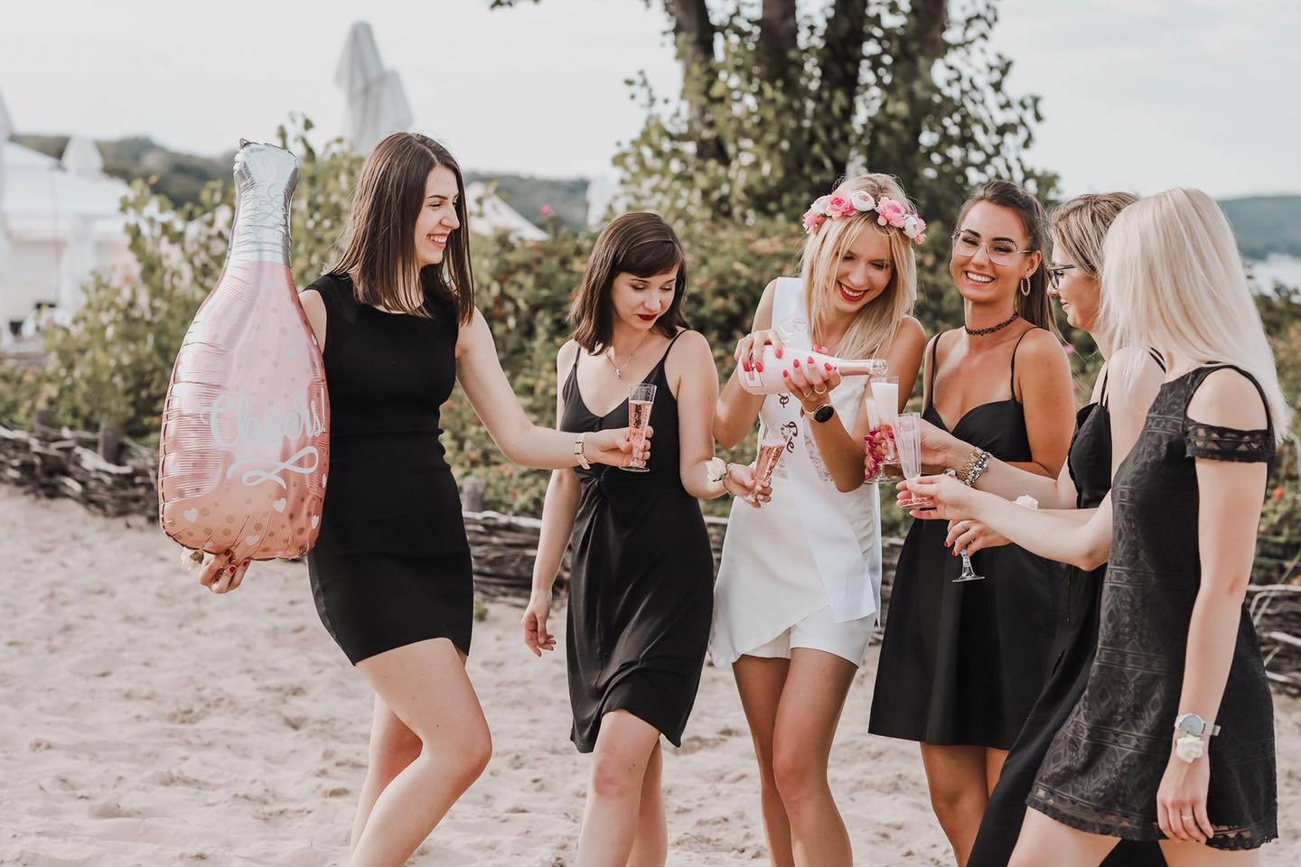 Grupa kobiet w sukienkach świętuje na plaży, jedna z nich w białej sukience i wianku nalewa różowe wino musujące do kieliszków, w tle balon w kształcie butelki szampana.
