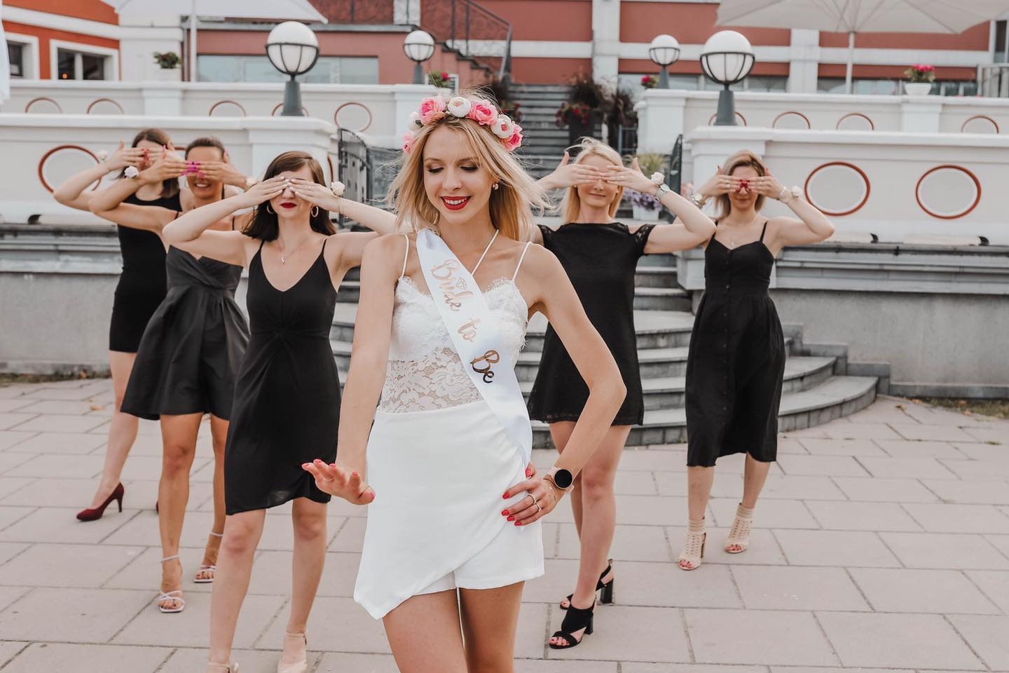 Przyszła panna młoda z kwiatowym wiankiem i szarfą 'Bride to Be' pozuje z druhnami, które zasłaniają oczy, na tle budynku z białymi balustradami i lampami.