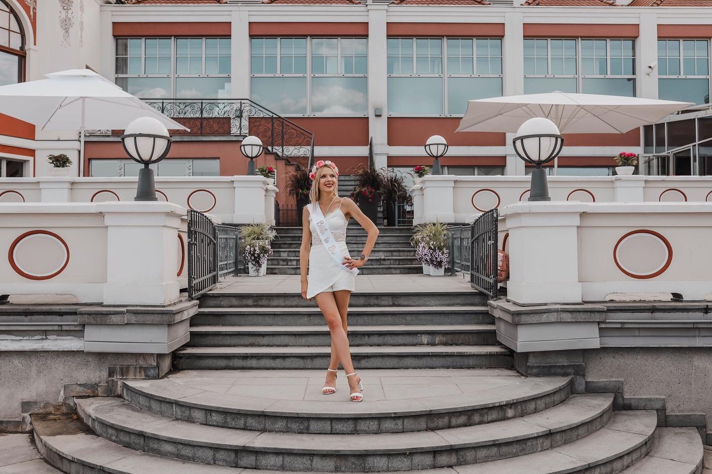 Uśmiechnięta kobieta w białym stroju z szarfą i wiankiem na głowie, pozuje na schodach przed budynkiem z klasyczną architekturą.