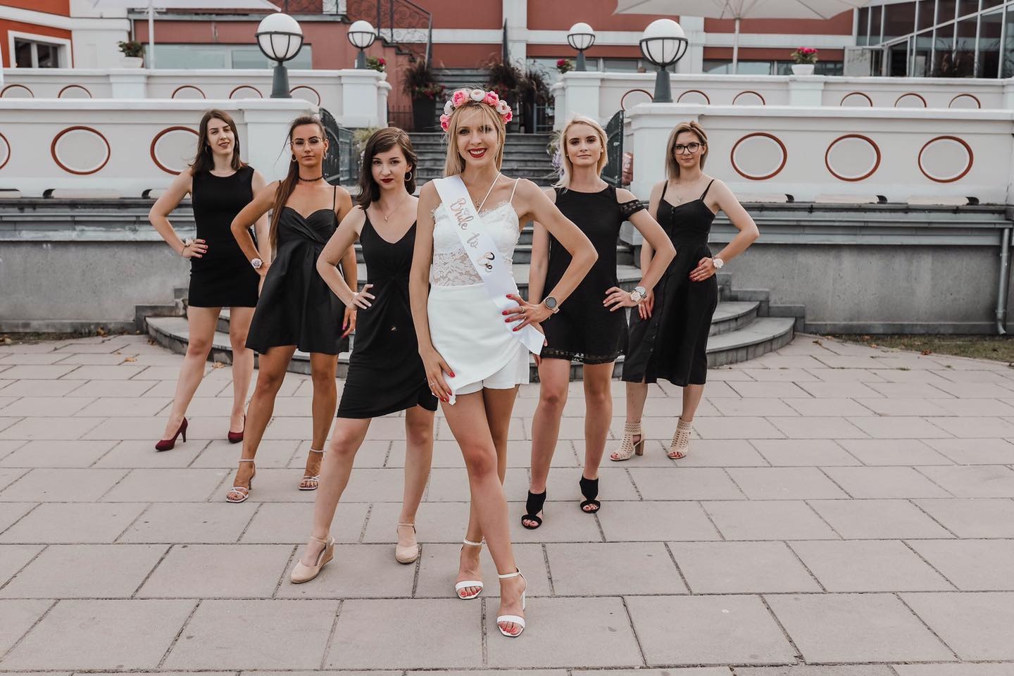 Panna młoda w koronie z kwiatów i szarfie 'Bride to be' pozuje z druhnami w czarnych sukienkach na tle budynku z białymi balustradami i schodami.