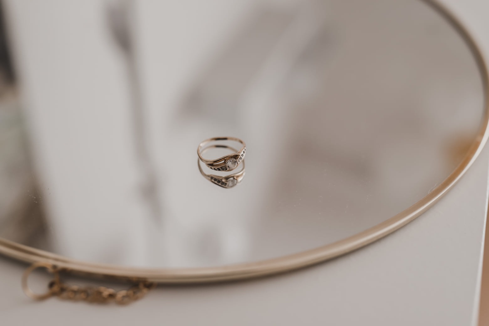 Złoty pierścionek z cyrkoniami na okrągłym lustrze z delikatnym odbiciem, obok fragment złotego łańcuszka.