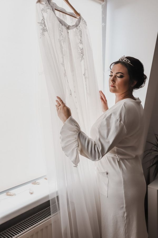 Panna młoda w białym szlafroku z tiarą, dotyka delikatnej sukni ślubnej wiszącej na wieszaku przy oknie.