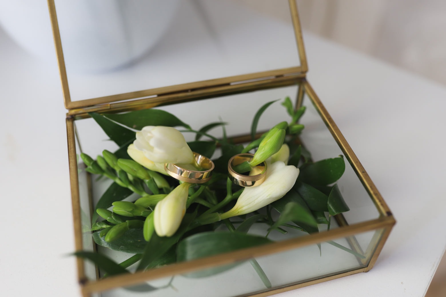 Dwa złote obrączki ślubne leżące na białych kwiatach frezji i zielonych liściach w szklanym, geometrycznym pudełku z mosiężnymi krawędziami.