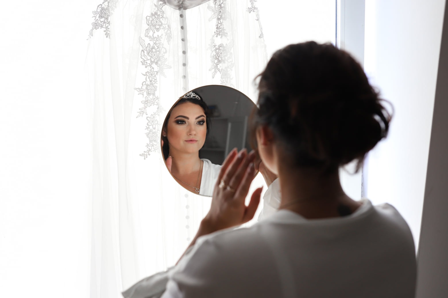 Kobieta z tiarą przegląda się w okrągłym lustrze, widoczna suknia ślubna z delikatnymi koronkowymi aplikacjami w tle.