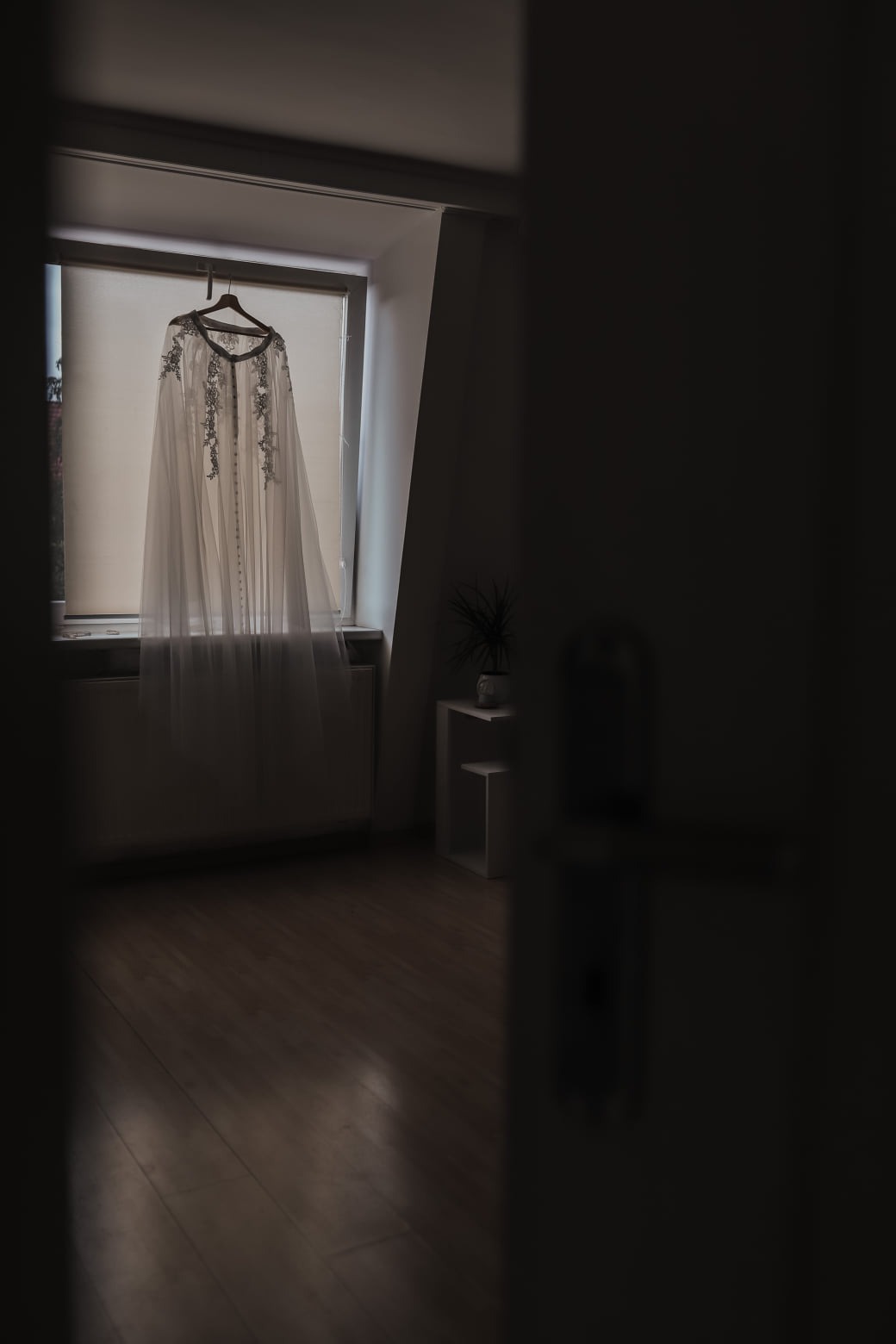 Delikatna, biała suknia z haftowanymi aplikacjami zawieszona na wieszaku przy oknie w słabo oświetlonym pokoju, widok zza drzwi.