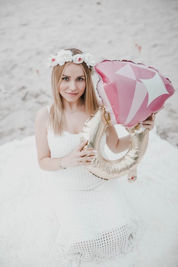 Blondynka w białej sukience i wianku z kwiatów trzyma balony w kształcie pierścionka zaręczynowego na tle piasku.