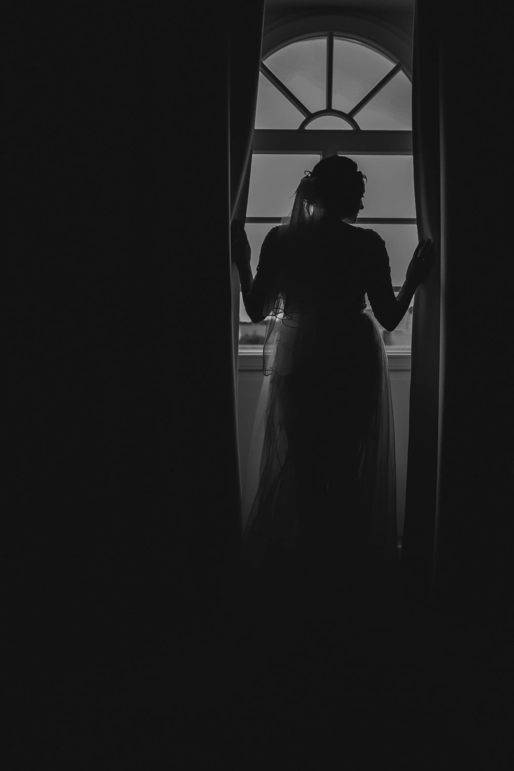 Sylwetka kobiety w sukni, stojącej tyłem przy oknie z rozchylonymi zasłonami, w czerni i bieli.