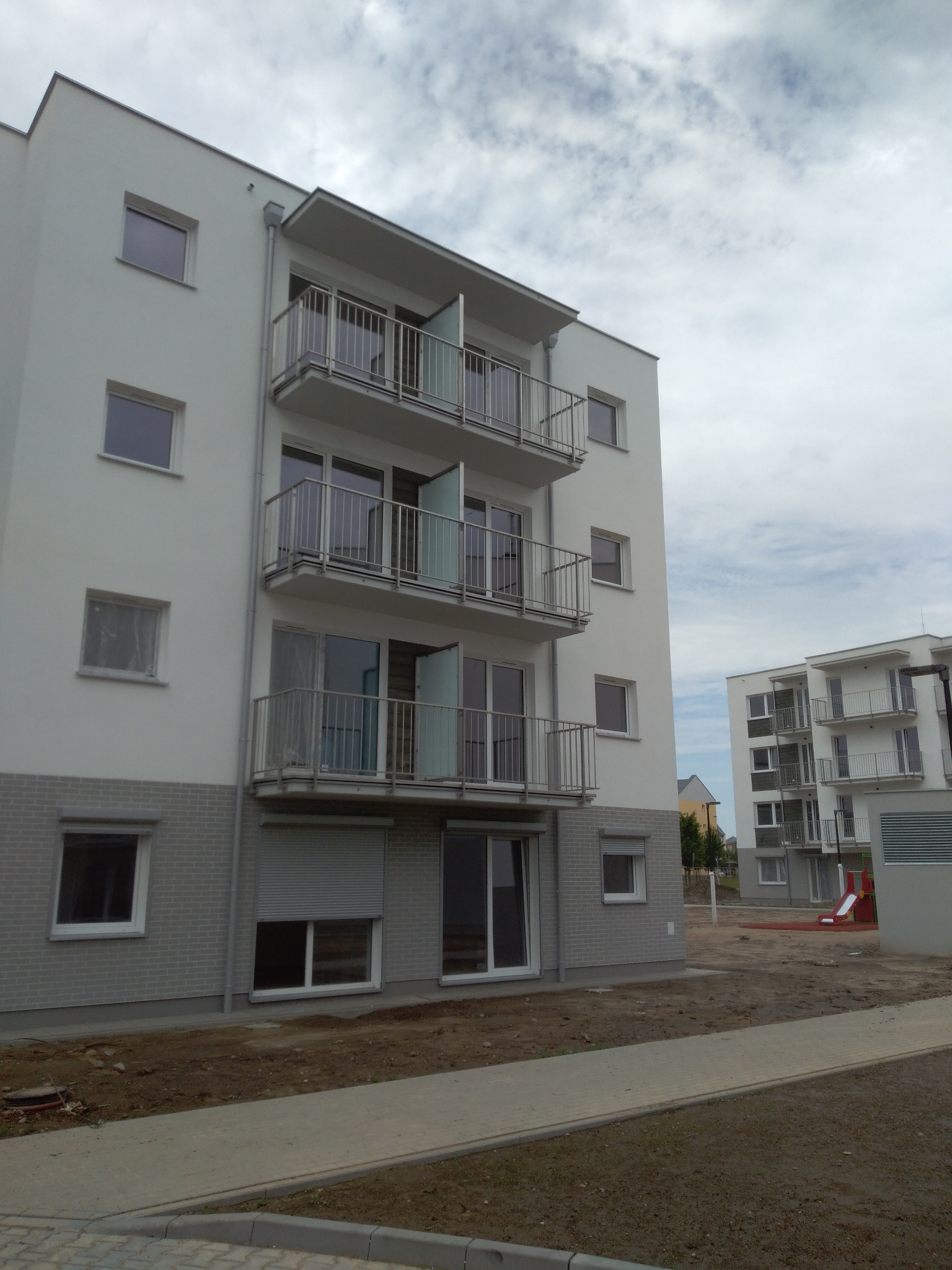 Nowoczesny budynek wielorodzinny z balkonami wyposażonymi w metalowe barierki, widok z poziomu chodnika, plac budowy w tle.
