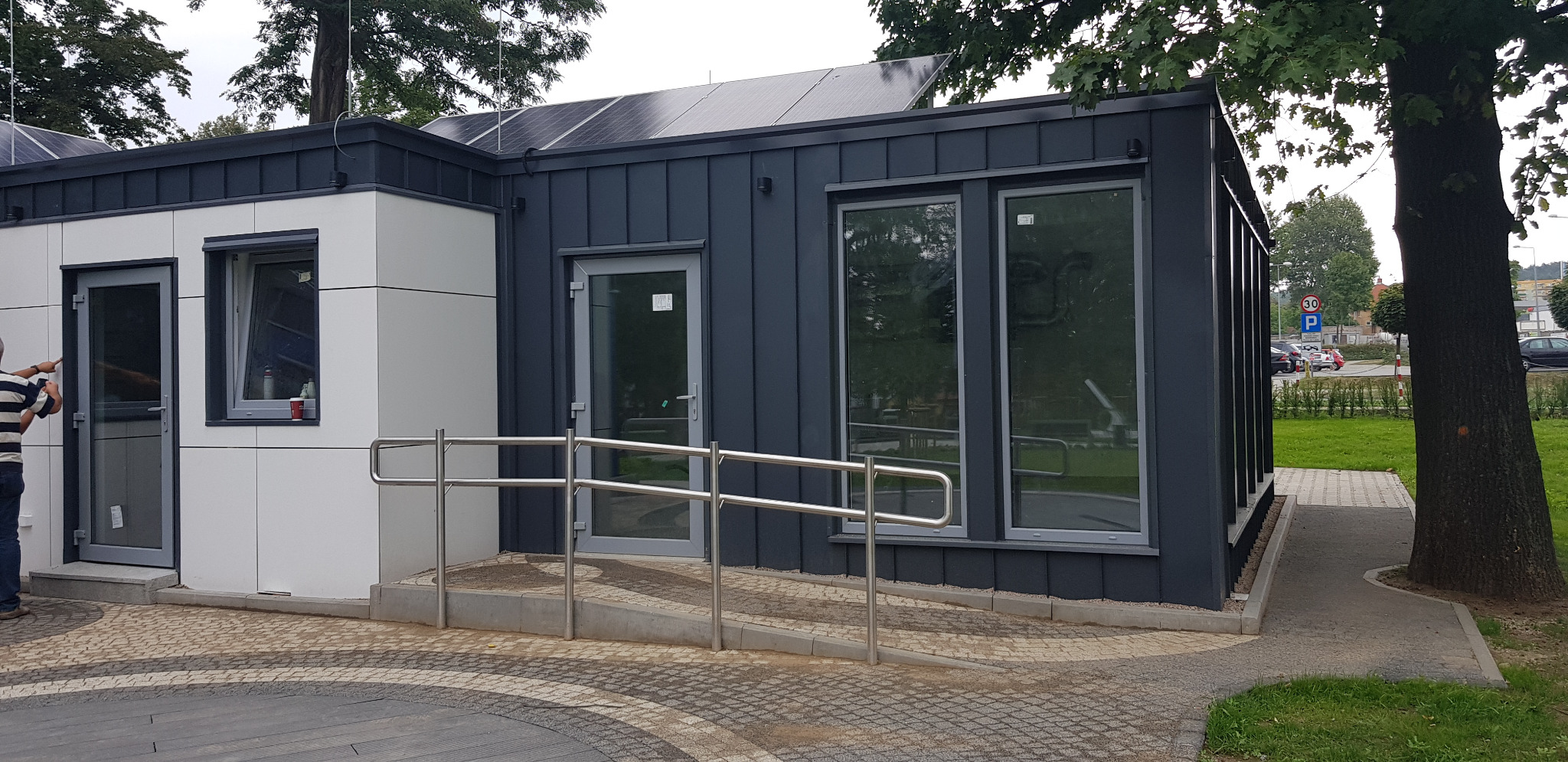 Nowoczesny pawilon z panelami słonecznymi na dachu, wyposażony w srebrne poręcze ułatwiające dostęp, z widocznym fragmentem osoby w paski po lewej stronie.