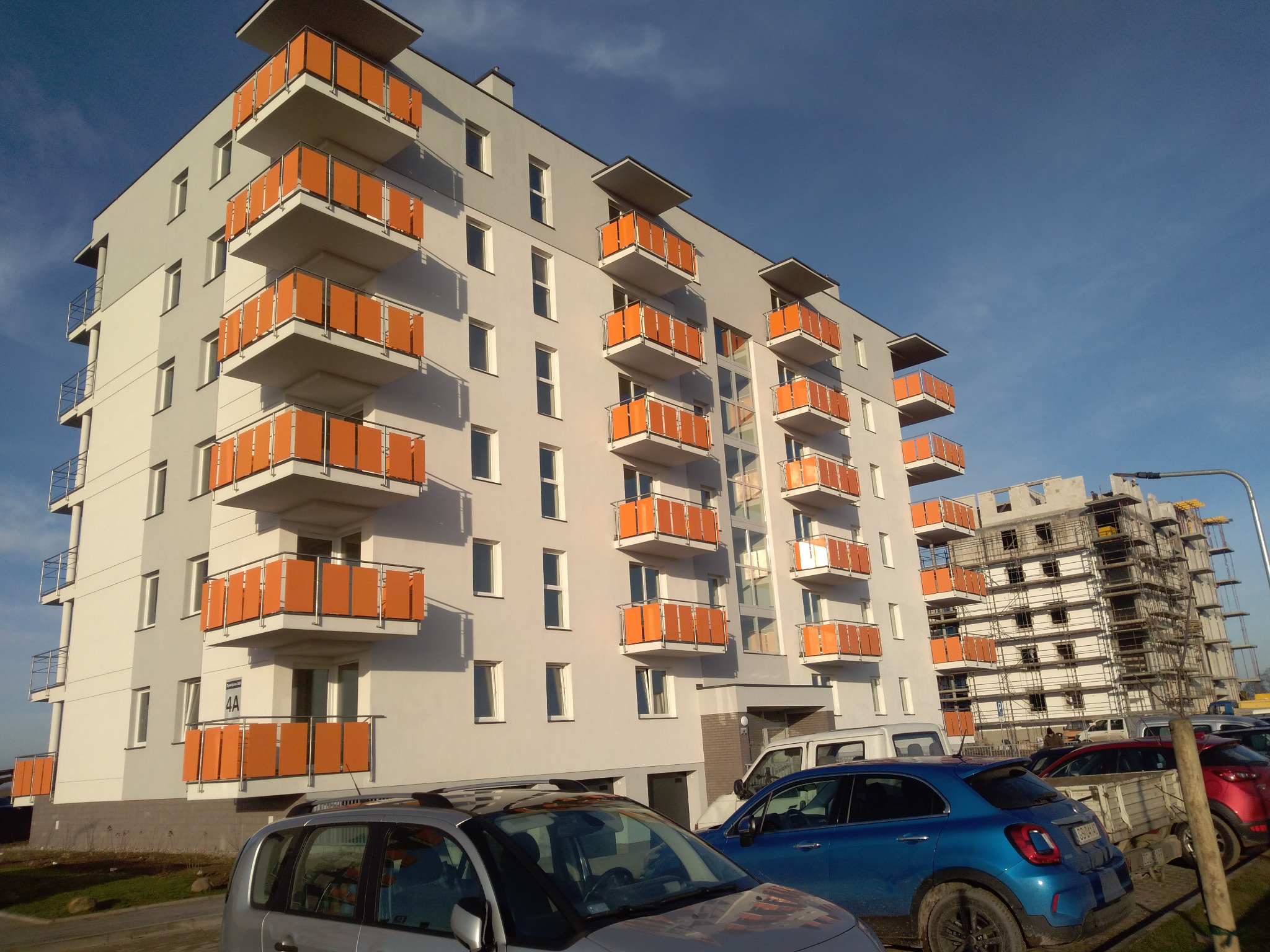 Nowoczesny budynek mieszkalny z balkonami wyposażonymi w pomarańczowe panele, widoczne samochody na parkingu i budowa innego budynku w tle.