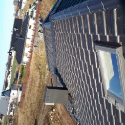 Nowy dach z ciemnej dachówki ceramicznej z oknem dachowym i kominem wentylacyjnym, widok z góry na tle domów jednorodzinnych.