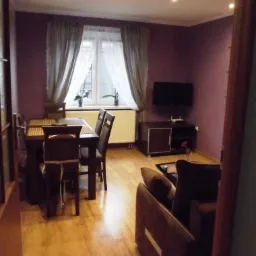 Salon z ciemnymi meblami: stół z krzesłami, brązowa sofa i szafka pod telewizorem. Sufit z punktowym oświetleniem.