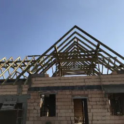 Surowy stan domu jednorodzinnego w trakcie budowy, widoczne ściany z cegły dziurawki i drewniana konstrukcja dachu pod błękitnym niebem.