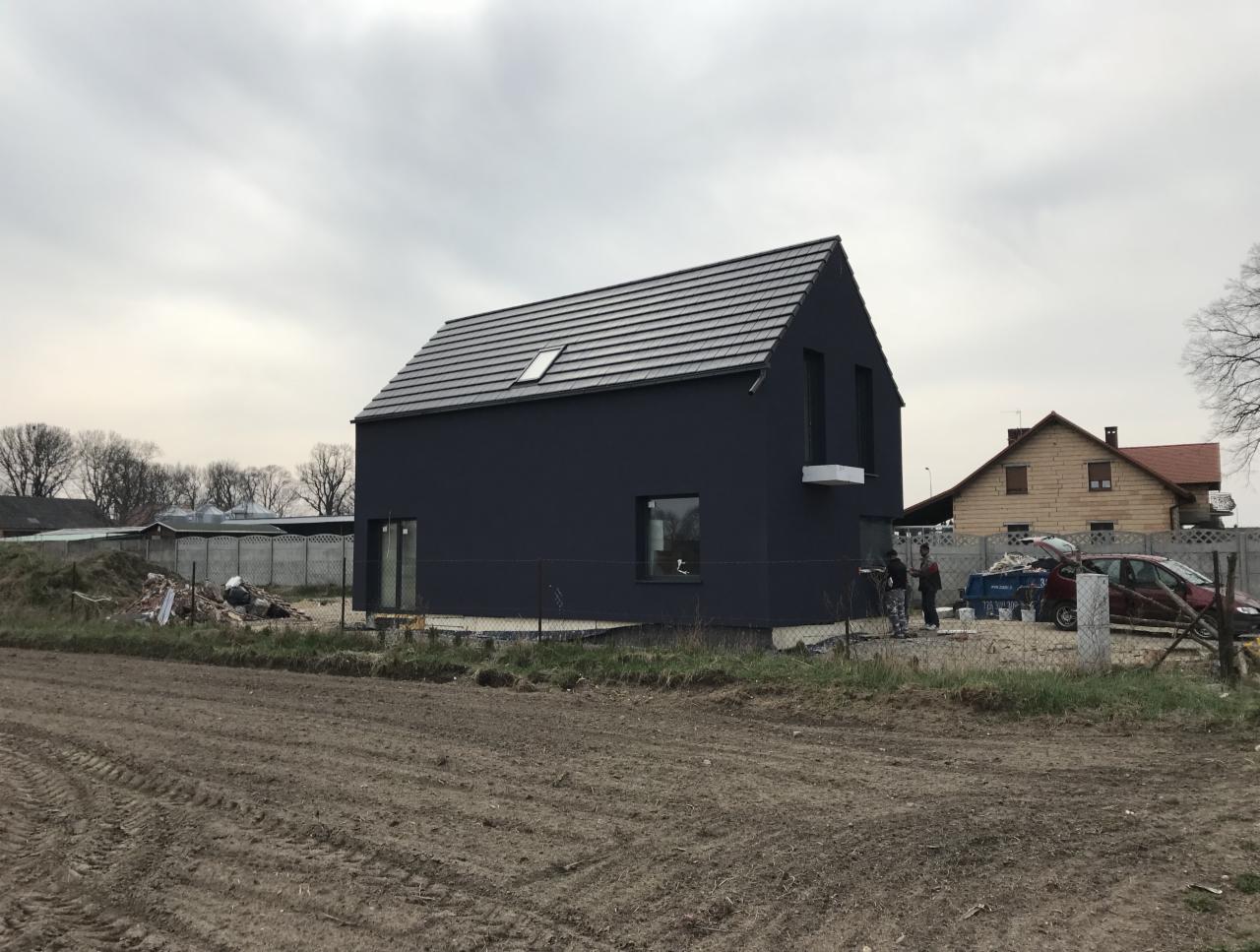 Nowoczesny, ciemny dom jednorodzinny z szarą dachówką i niewielkim oknem dachowym, widoczny z perspektywy gruntu, z pracownikami przy samochodzie w tle. Teren wokół domu częściowo zagospodarowany...