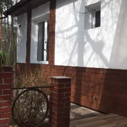 Elewacja domu z białym tynkiem i brązową boazerią, widoczne okna, ceglane słupki i drewnopodobna płytka na schodach wejściowych, cień drzew na ścianie.