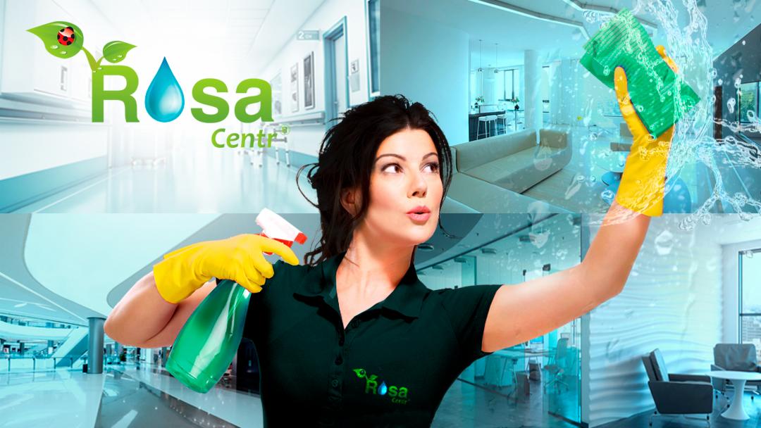 Kobieta w żółtych rękawicach spryskuje i czyści szybę zieloną gąbką, w tle widoczny hol biurowy i wnętrze apartamentu, na koszulce widoczne logo firmy sprzątającej.