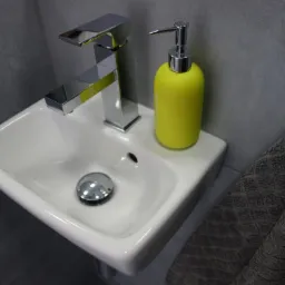 Nowoczesna, mała umywalka z chromowaną baterią i żółtym dozownikiem mydła, szara płytka ścienna i ręcznik z fakturą rombów.