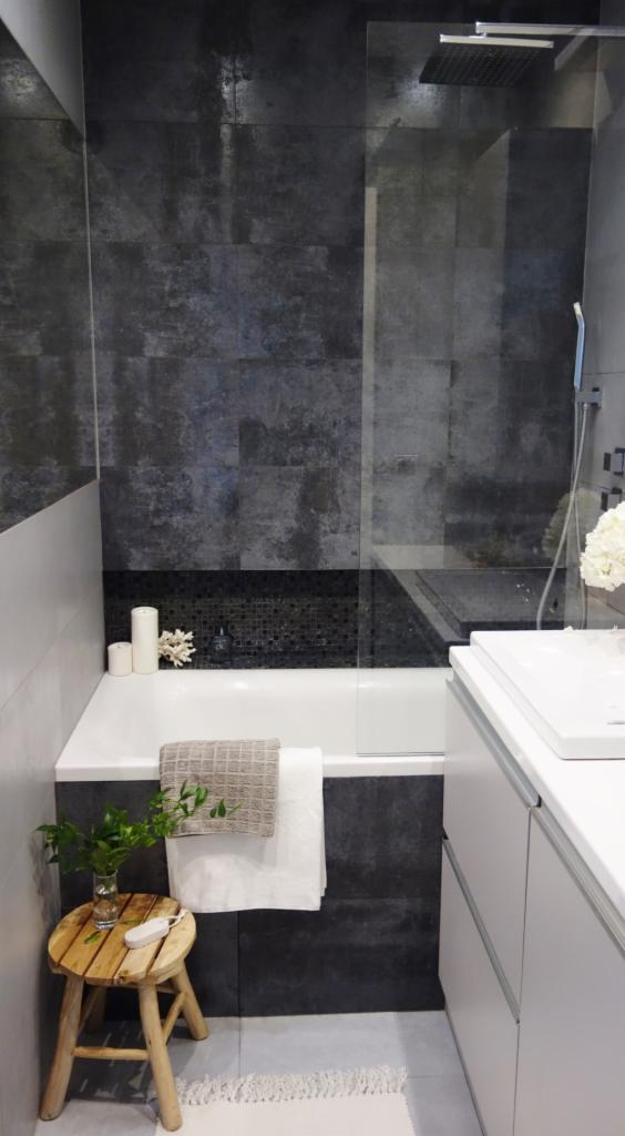 Szara łazienka z wanną, kabiną prysznicową typu walk-in, drewnianym stołkiem z rośliną i ręcznikami.