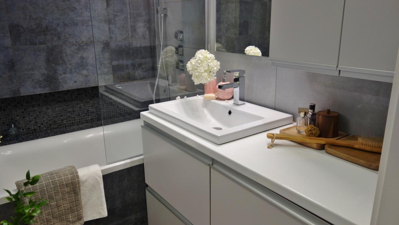 Nowoczesna łazienka z białą szafką umywalkową, chromowaną baterią, szarą mozaiką nad wanną i drewnianymi akcesoriami na blacie.