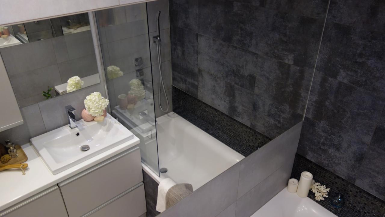 Nowoczesna łazienka z wanną za szklaną ścianką, umywalką z szafką w odcieniach szarości i białymi akcentami dekoracyjnymi, takimi jak kwiaty i świece.