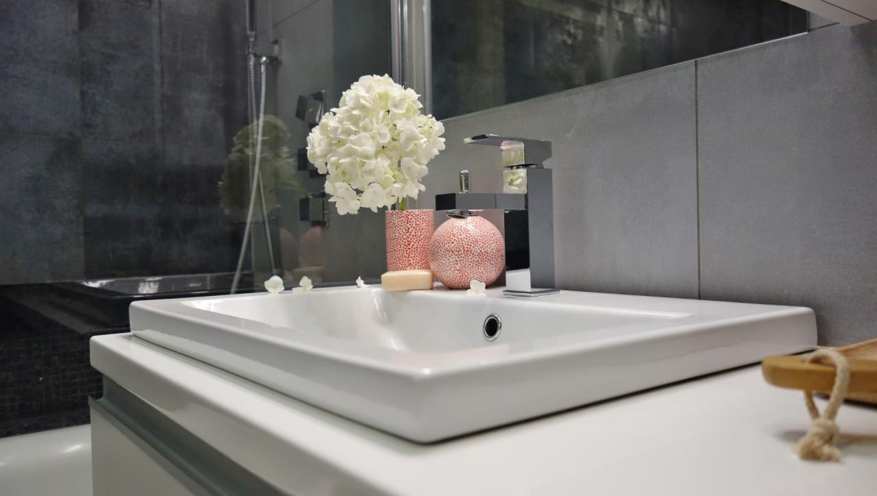 Nowoczesna, minimalistyczna umywalka w łazience z chromowaną baterią, dekoracyjnymi pojemnikami w kropki, mydłem i bukietem białych kwiatów na tle szarych płytek.