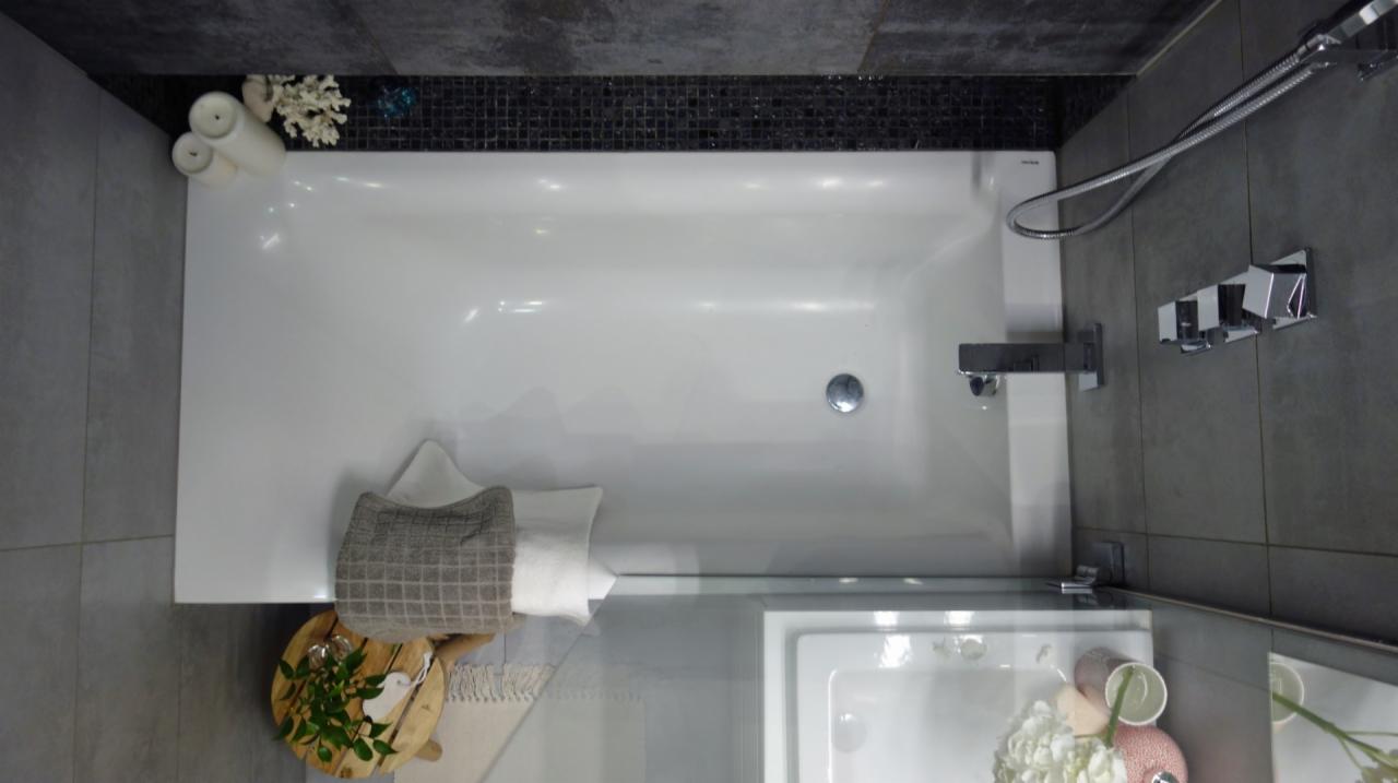 Nowoczesna łazienka z białą wanną, szarą glazurą i dekoracyjnymi elementami: świece, koralowce i drewniana taca z zielonymi liśćmi.