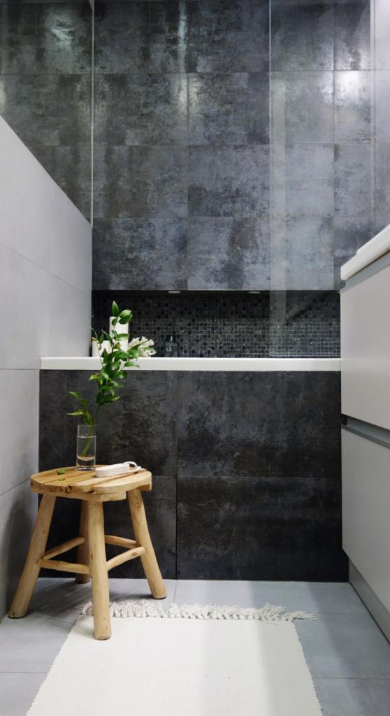 Nowoczesna łazienka z ciemnymi, teksturowanymi płytkami, drewnianym stołkiem z wazonem i białym dywanikiem.