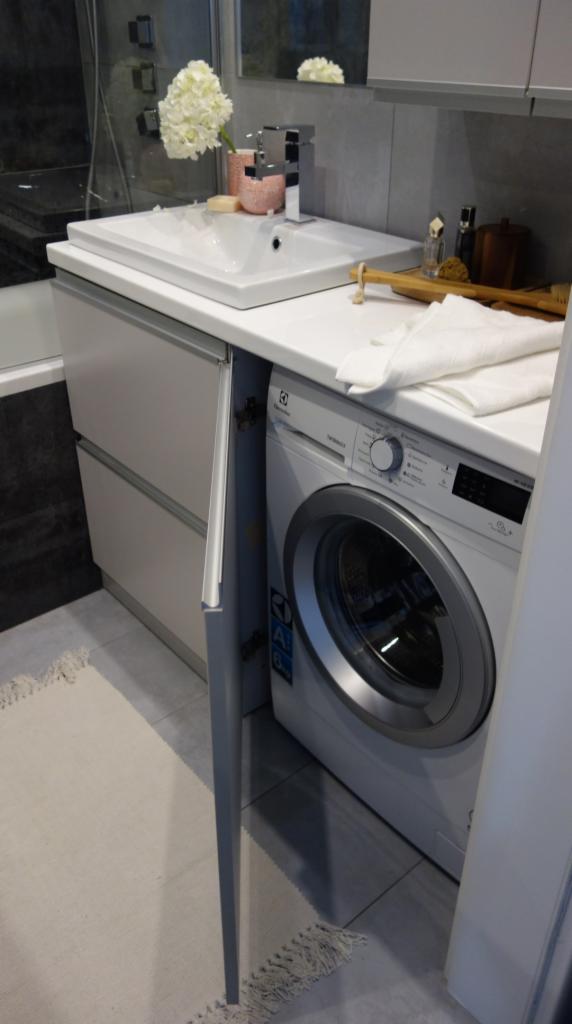 Nowoczesna łazienka w szarościach z pralką Electrolux ukrytą pod blatem umywalki, otwarte drzwiczki szafki, biały ręcznik, kwiaty w wazonie i akcesoria na blacie.