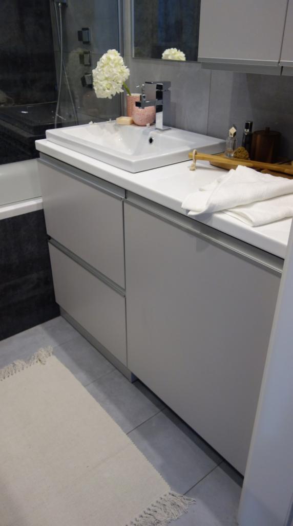 Nowoczesna łazienka w odcieniach szarości z białą umywalką nablatową, kwiatami w różowym wazonie i minimalistyczną szafką pod umywalką z szufladami oraz dywanikiem z frędzlami.