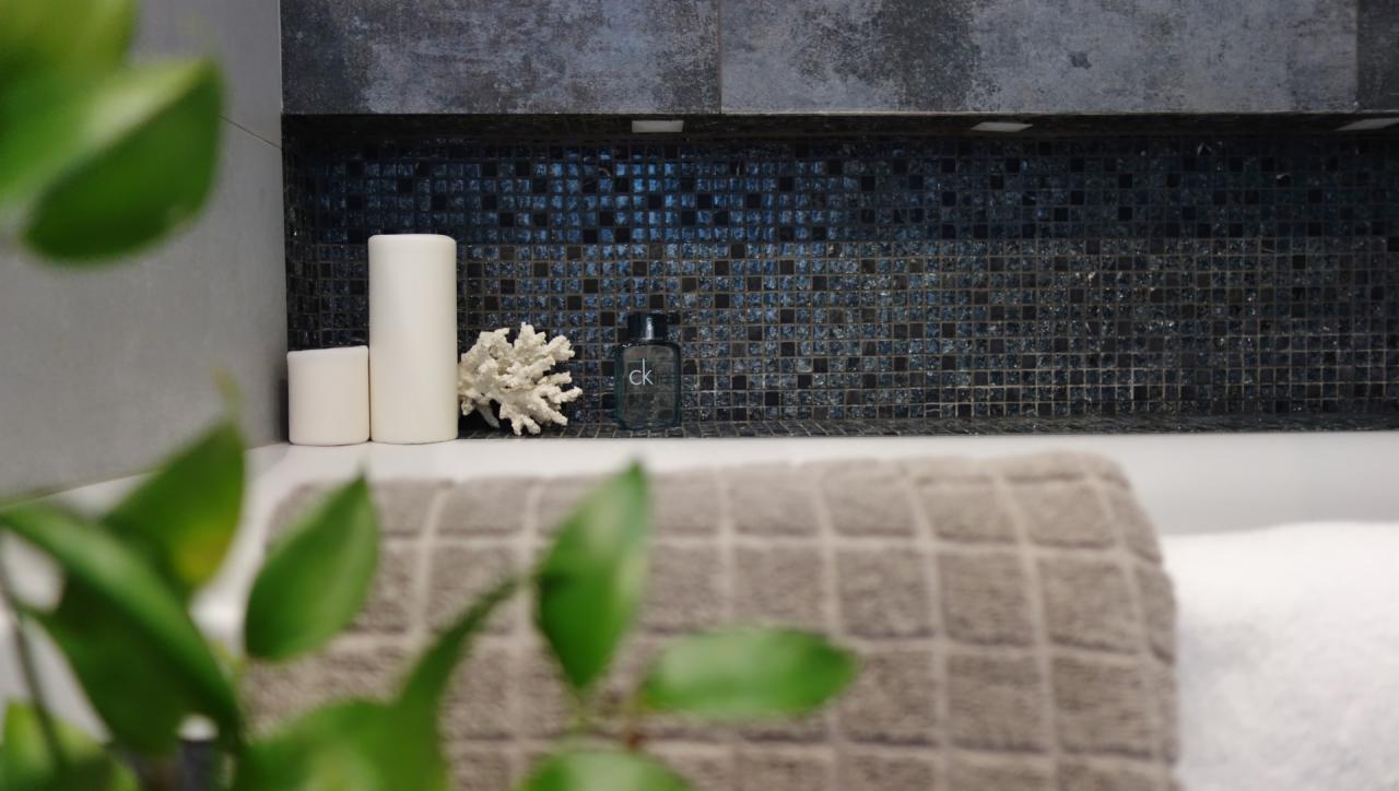 Nowoczesna łazienka z mozaiką, białymi świecami, koralem i butelką perfum CK, ręczniki w kratkę, roślina w tle.