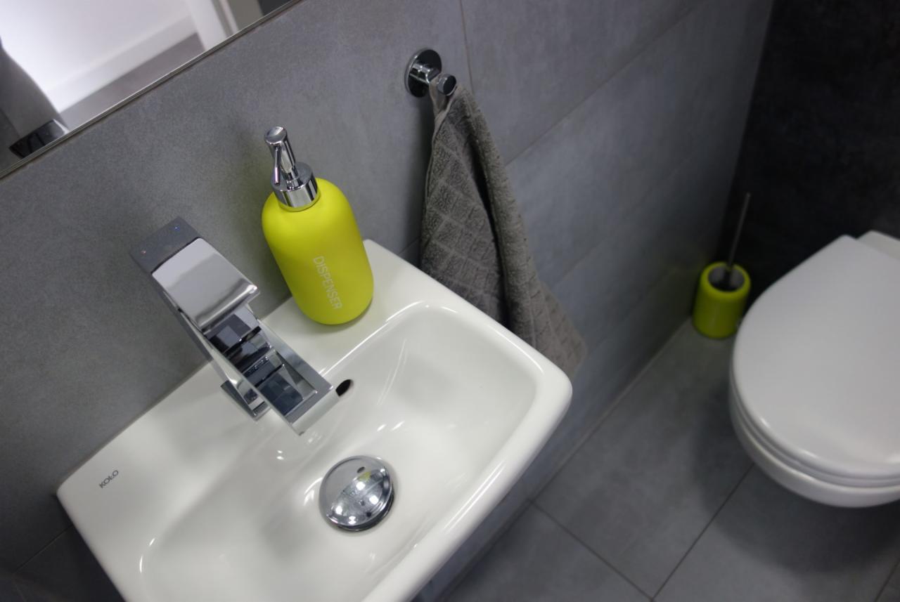 Nowoczesna, szara łazienka z białą umywalką KOLO, chromowaną baterią, żółtym dozownikiem mydła i szarym ręcznikiem na wieszaku, z toaletą w tle.