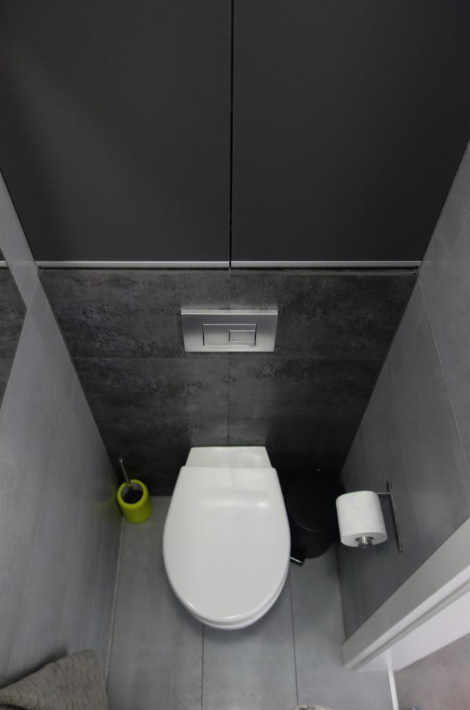 Nowoczesna toaleta w szarościach, widok z góry, z podwieszaną muszlą klozetową, przyciskiem spłukującym w kolorze stali, szarą szczotką do WC z żółtym uchwytem i rolką papieru toaletowego...