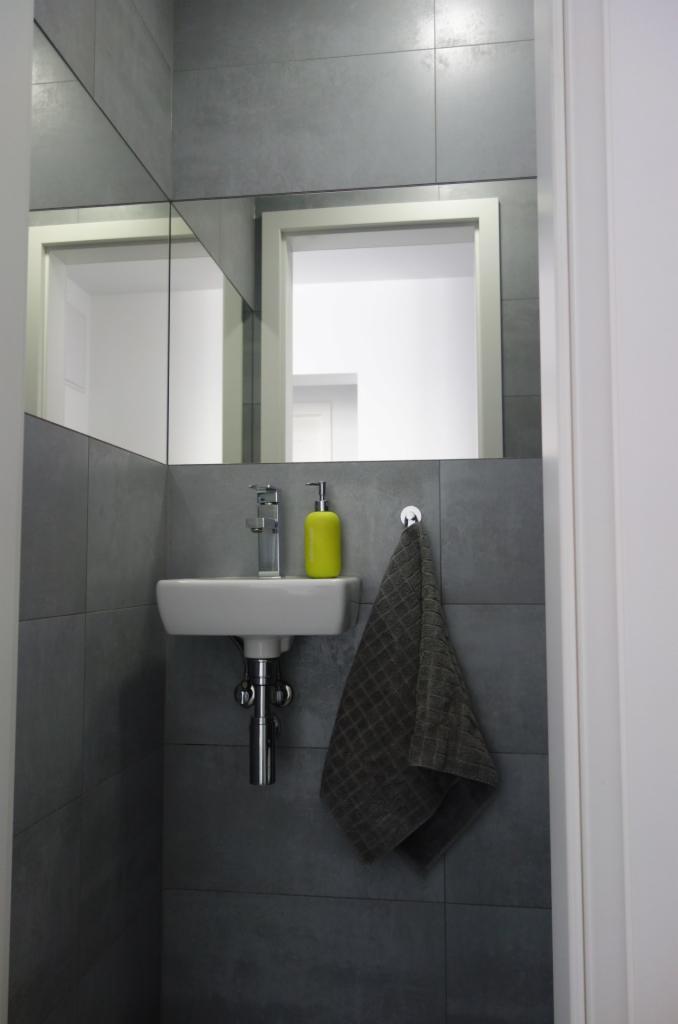 Mała łazienka z umywalką nablatową, baterią, żółtym dozownikiem mydła i szarą płytką na ścianach; lustro odbija wnętrze pomieszczenia.