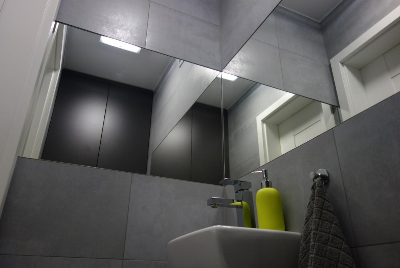 Nowoczesna łazienka z szarymi płytkami, lustrem składającym się z kilku części, białą umywalką, chromowaną baterią i żółtym dozownikiem mydła. W lustrze odbija się ciemnoszara szafka i białe drzwi.