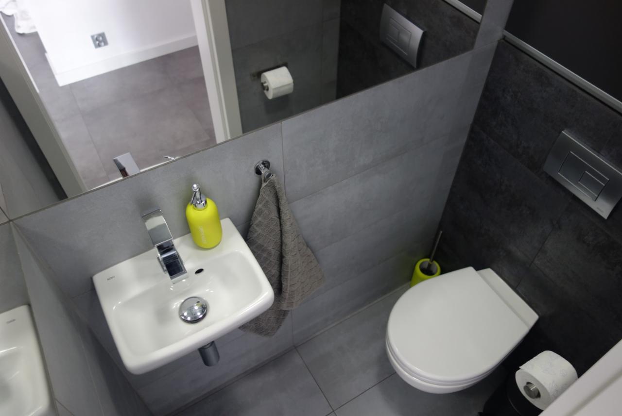 Nowoczesna łazienka z szarymi płytkami, białą umywalką i toaletą, akcentem kolorystycznym w postaci żółtego dozownika mydła i pojemnika na szczotkę toaletową.