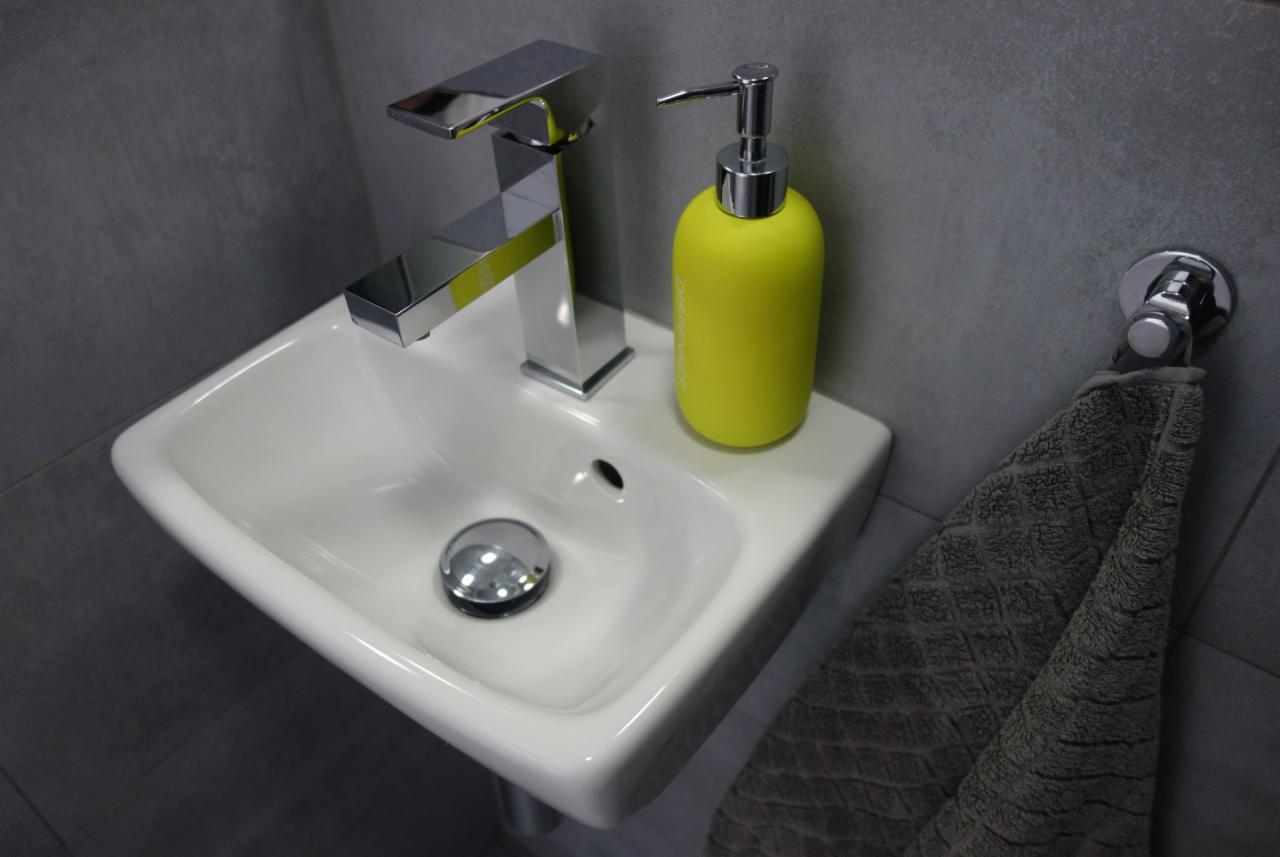 Nowoczesna, mała umywalka z chromowaną baterią i żółtym dozownikiem mydła, szara płytka ścienna i ręcznik z fakturą rombów.