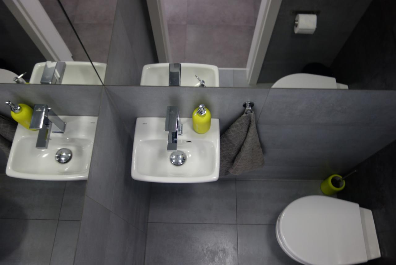 Umywalka nablatowa z chromowaną baterią, żółtym dozownikiem mydła i szarą ściereczką wiszącą na haczyku, widok z góry, szare płytki na ścianach i podłodze, toaleta w tle