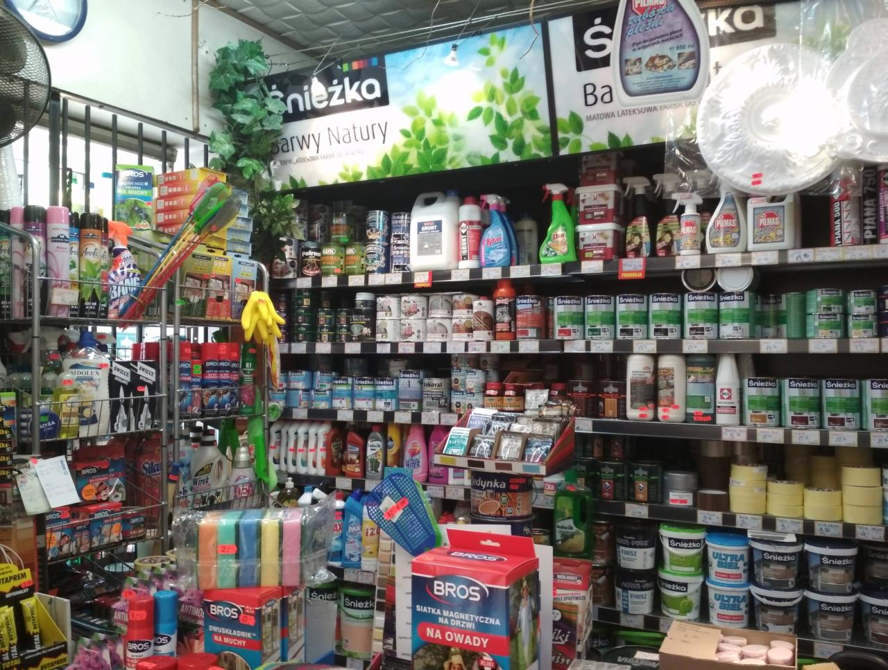 Wnętrze sklepu z artykułami budowlanymi, regały wypełnione farbami, lakierami, środkami czystości i akcesoriami, widoczny wentylator sufitowy i reklama marki Śnieżka.