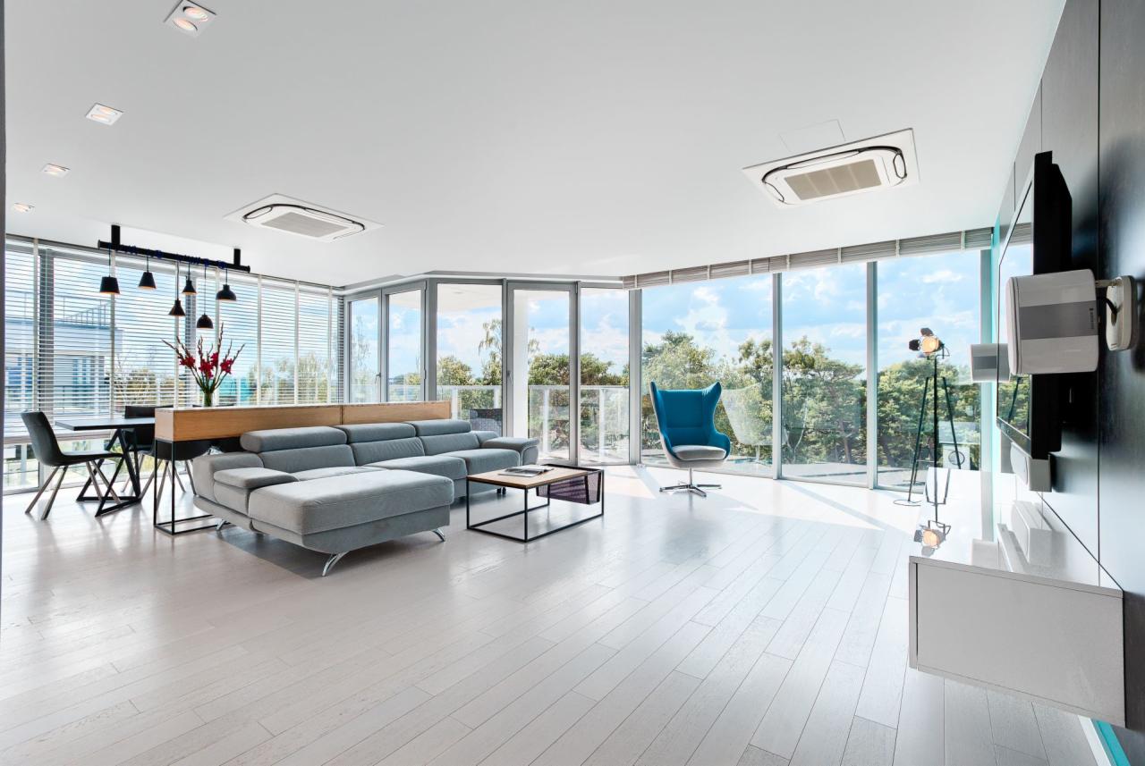 Jasne, przestronne wnętrze z dużymi oknami, szarą sofą narożną, turkusowym fotelem i drewnianym stołem jadalnym z czarnymi krzesłami, oświetlone wiszącymi lampami i światłem dziennym.