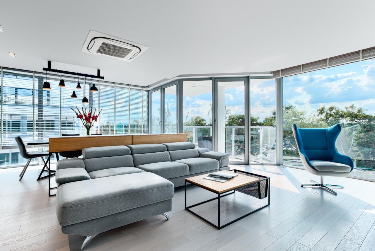 Jasne, przestronne wnętrze salonu z szarą sofą narożną, nowoczesnym stolikiem kawowym i turkusowym fotelem na tle panoramicznych okien z widokiem na zieleń.