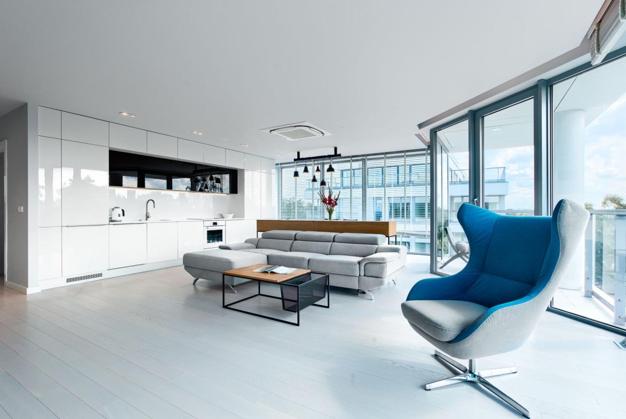 Jasne, nowoczesne wnętrze z białą, błyszczącą zabudową kuchenną, szarą sofą narożną, stolikiem kawowym z gazetnikiem i fotelem w kolorze szaro-niebieskim na tle przeszklonej ściany z widokiem...