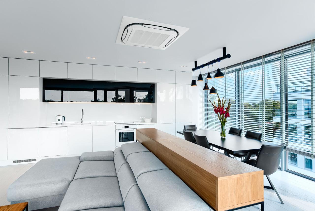 Nowoczesna kuchnia z jadalnią w jasnych kolorach, z białymi błyszczącymi szafkami, czarnymi akcentami i szarym narożnikiem z drewnianym panelem oddzielającym przestrzeń.