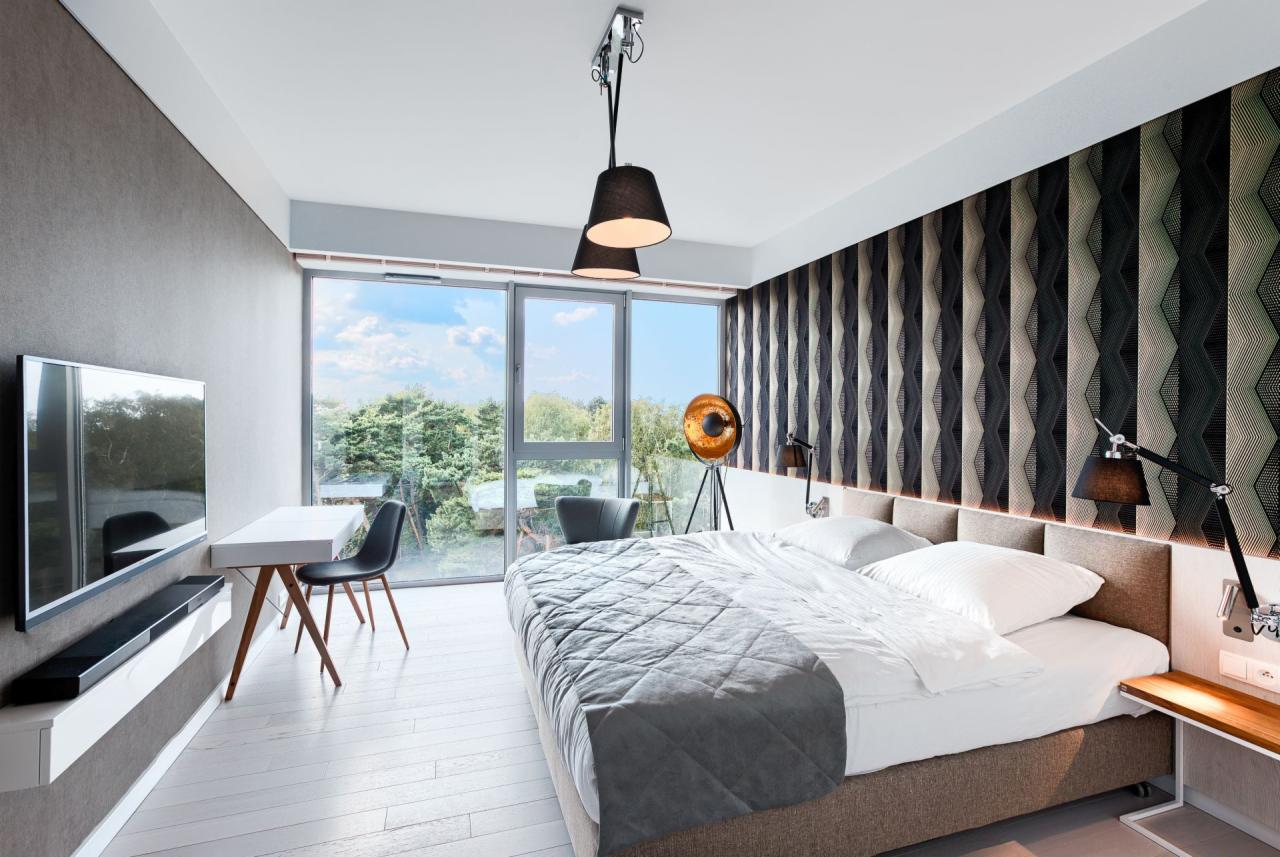 Nowoczesna sypialnia z dużym oknem, biurkiem i wzorzystą tapetą za łóżkiem. Widoczne lampy wiszące i stojące, oraz telewizor na ścianie.